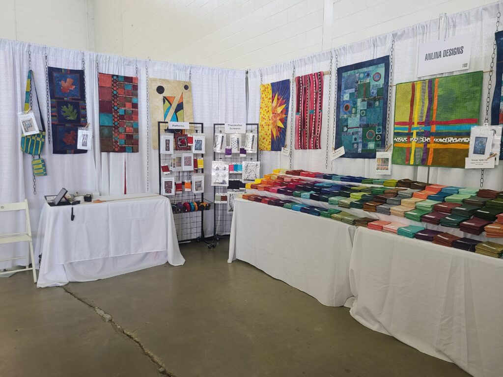 a booth with 7 quilts and a purse hanging on white curtained panels, 3 tables, 2 of which have a rainbow of fabric squares on them; 2 grid wall panels with patterns, trim and screens sit between the tables in the back corner.